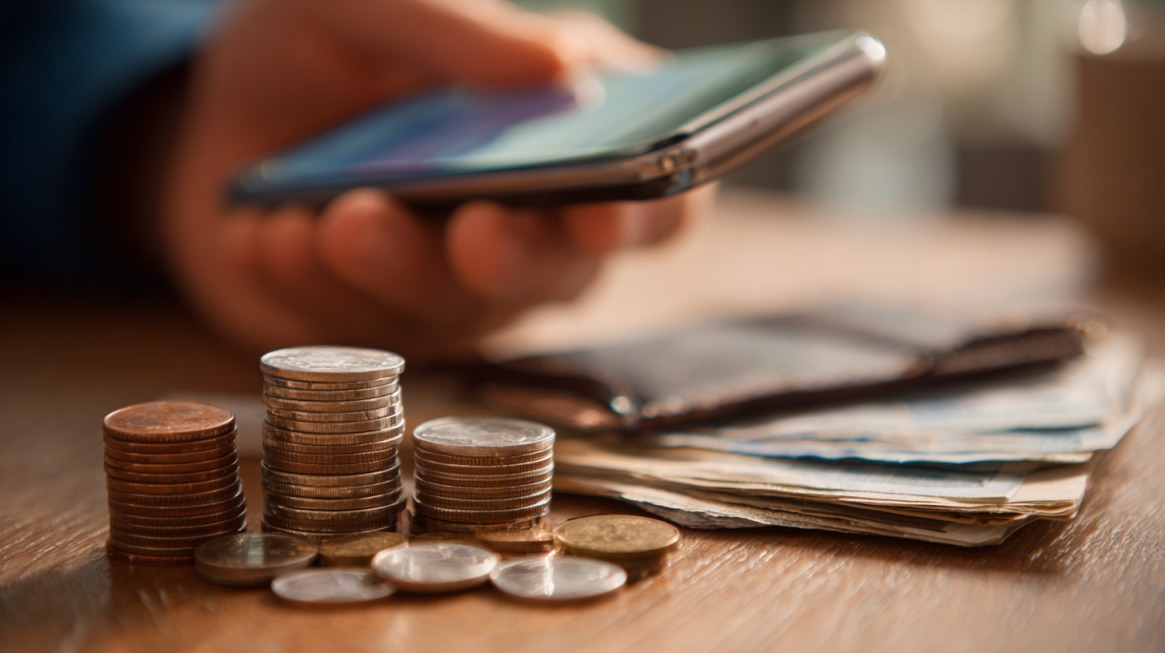 Pametni telefon na stolu pored naslaganih kovanica i novčanica, simbolizujući digitalno upravljanje ličnim finansijama putem mobilnih aplikacija